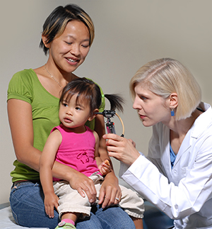 Toddler sitting on woman's lap while doctor examines her ear with otoscope.